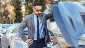 A man in a jacket and tie standing over a car with an open hood clearly having car trouble