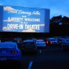 Rows of cars at a drive-in movie theater at dusk, trees in the background.